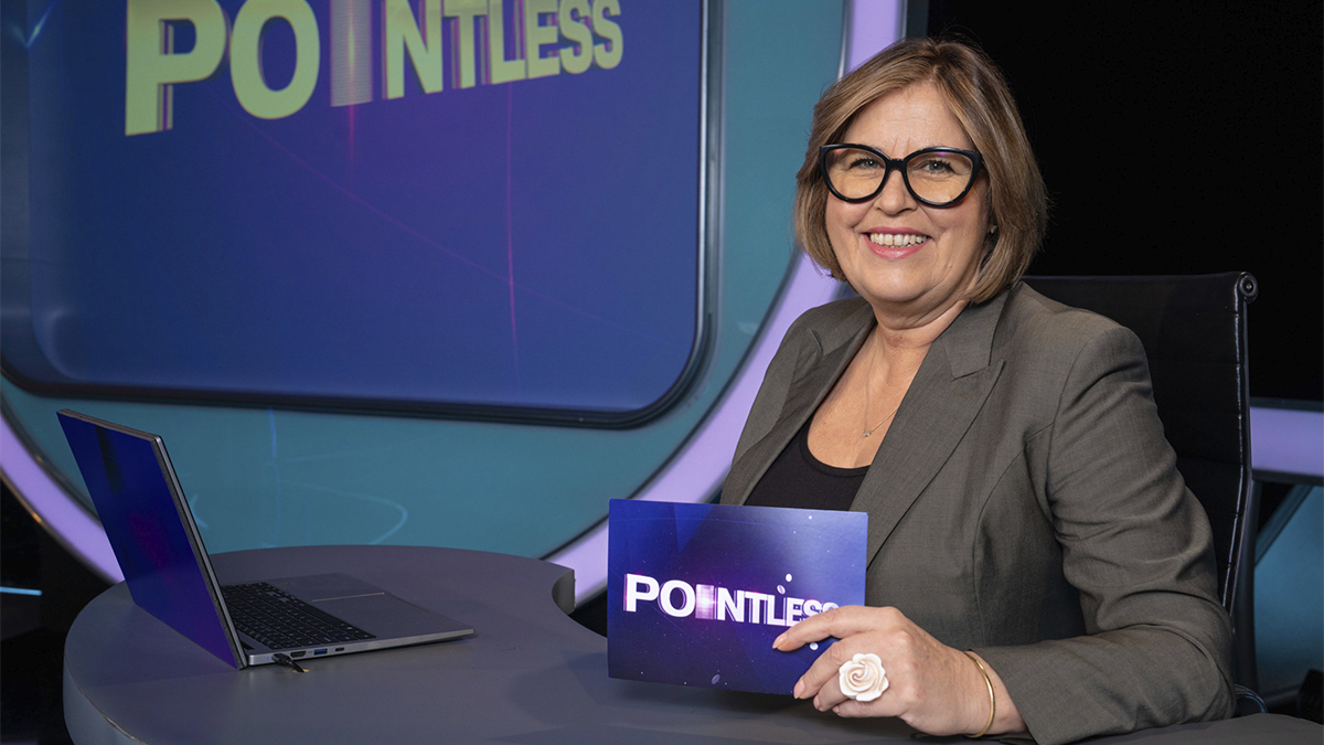 Liza Tarbuck wearing a grey suit and glasses sitting behind the Pointless desk